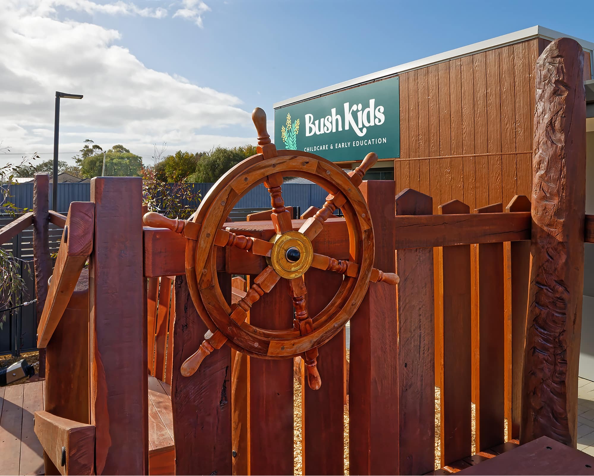 A large wooden ship’s wheel is mounted on a wooden play structure outdoors, with the Bush Kids Child Care & Early Education building visible in the background.